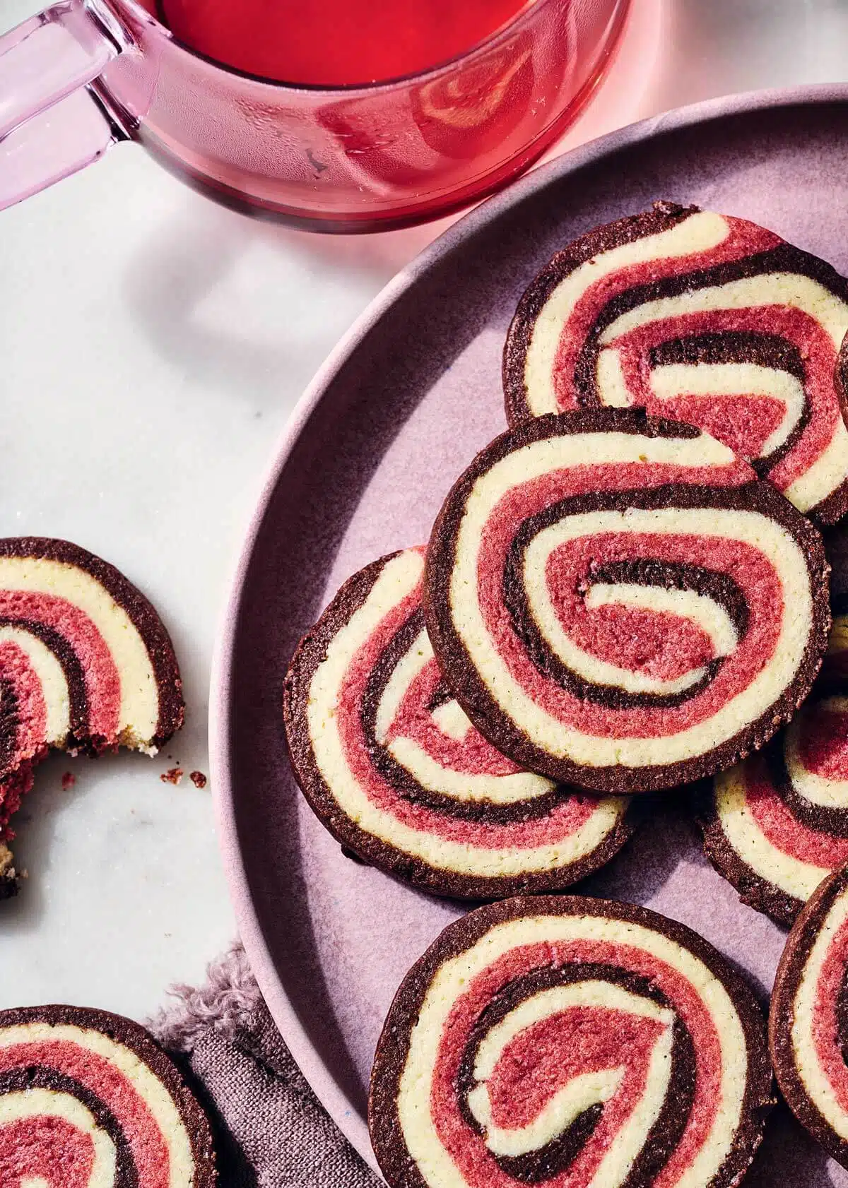 Neapolitan Pinwheel Cookies. Red and cream-colored pinwheel cookies on a plate next to a glass teapot.