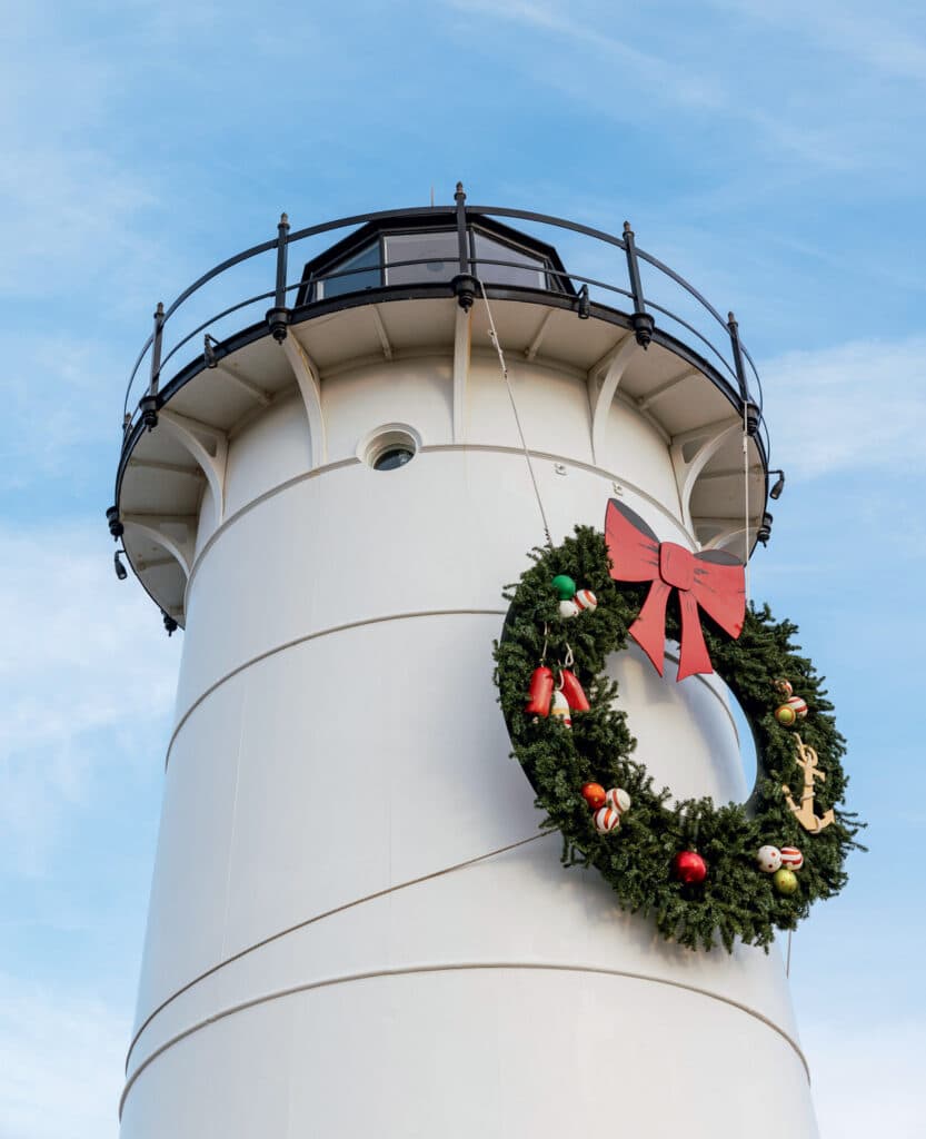 A white lighthouse adorned with a large Christmas wreath featuring a red bow and nautical ornaments.