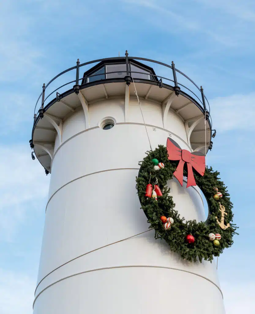 A white lighthouse adorned with a large Christmas wreath featuring a red bow and nautical ornaments.
