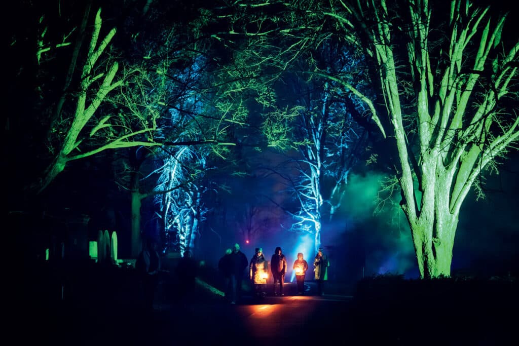 A group of people carrying lanterns walk along a path lined with trees illuminated by green and blue lights.