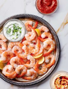 Jumbo Shrimp with Dipping Sauces. A silver platter of cooked shrimp with lemon wedges, a bowl of herb dip, red sauce, and hummus.