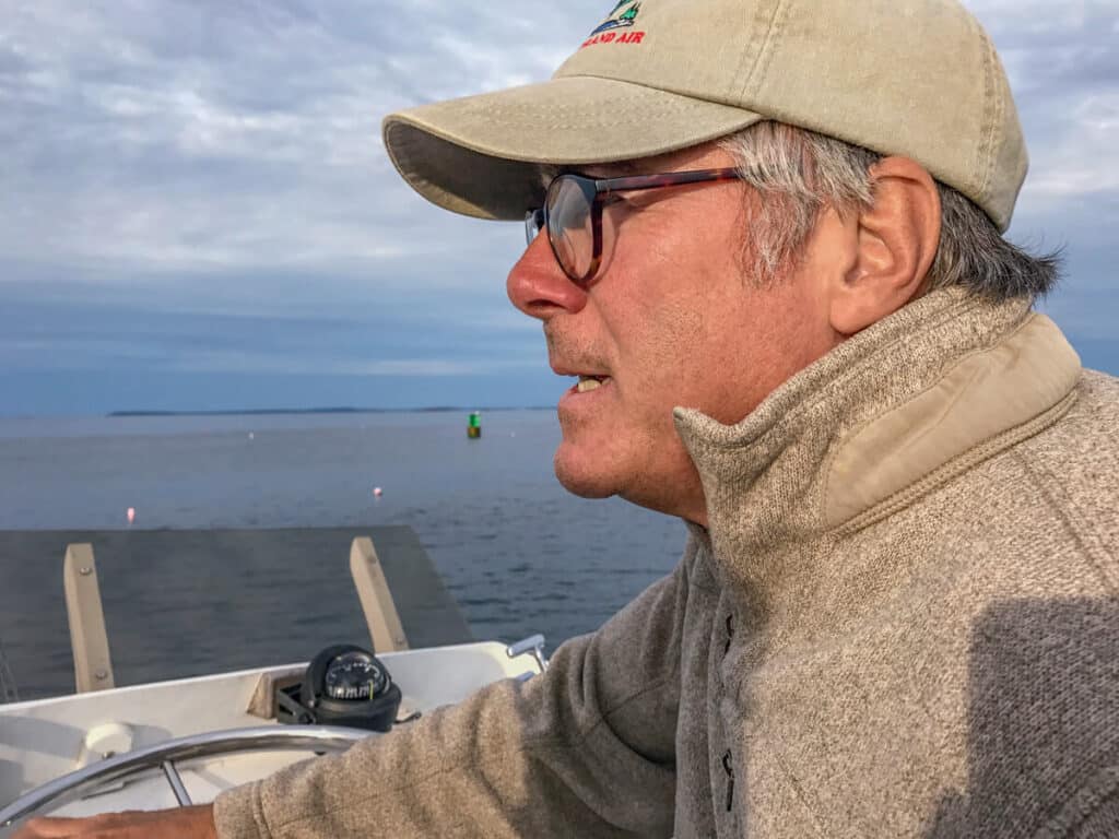 A man in a cap and glasses steering a boat on the water.