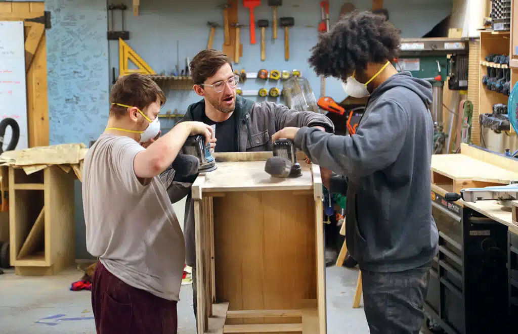 Three people working together to sand a wooden furniture frame in a workshop.
