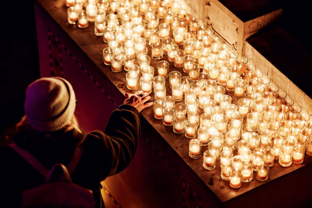 A person reaching toward rows of glowing votive candles on a ledge.