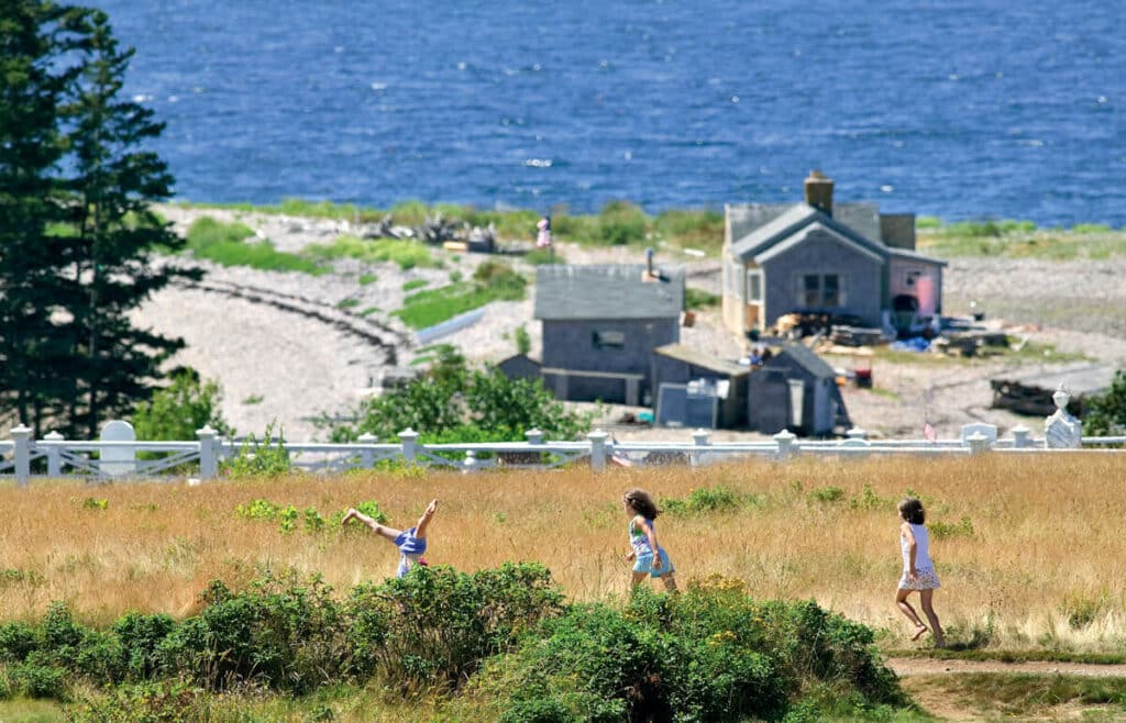 Three children play in a grassy field above a coastal beach with houses.