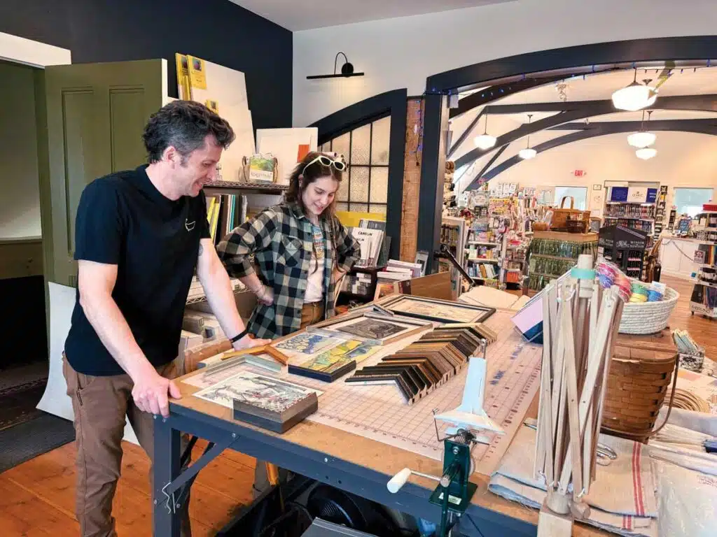 A man and woman examine framed artwork and matting samples on a large workbench in an art supply store.