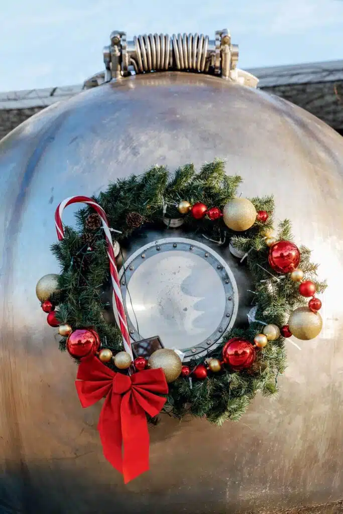 Christmas wreath with red and gold ornaments and a candy cane hanging on a large stainless steel industrial tank.