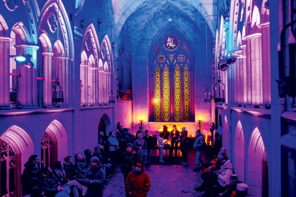 A group of people standing in a gothic-style hall illuminated by purple and yellow lights.