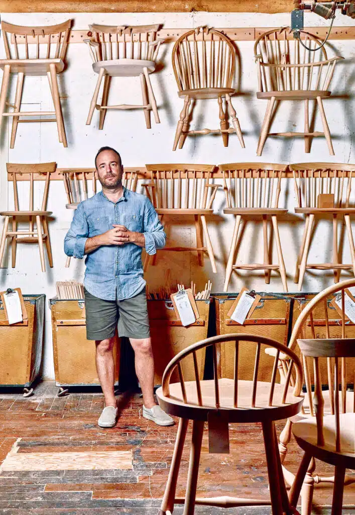 A man standing in a workshop surrounded by unfinished wooden chairs hanging on the wall and arranged on the floor.