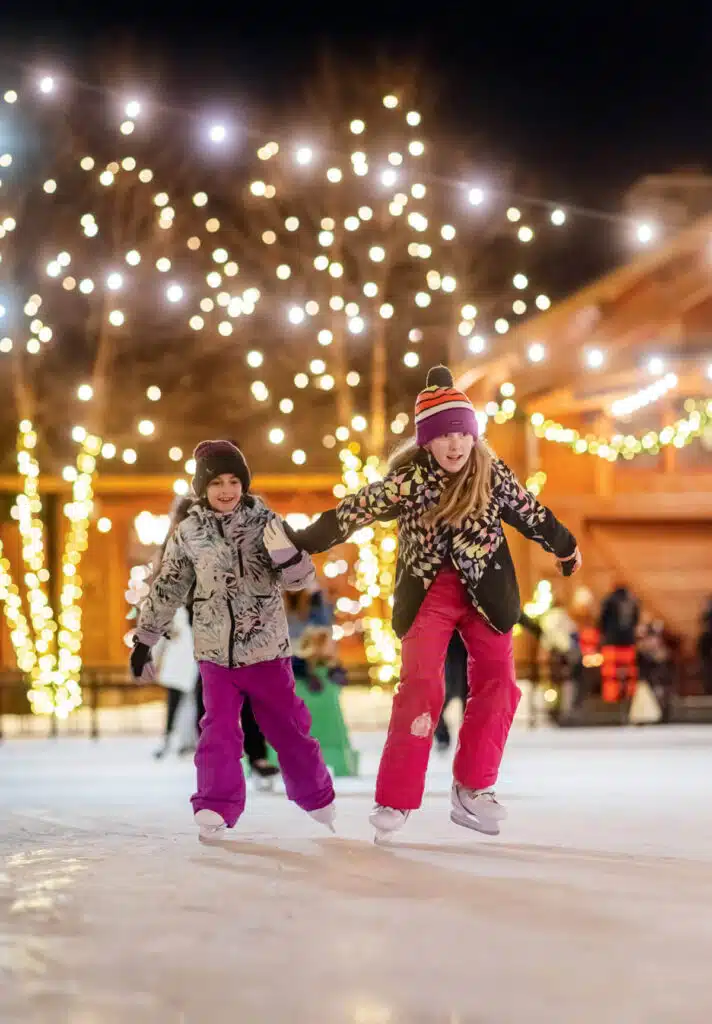 Two children ice skating together in front of illuminated trees.