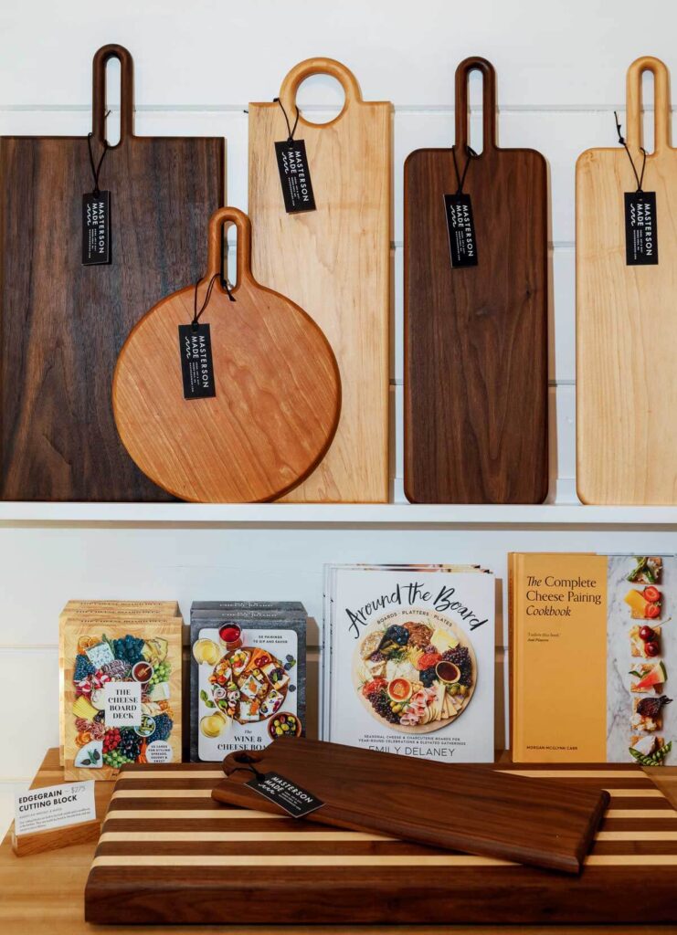 Wooden serving boards hanging on a wall above shelves displaying charcuterie cookbooks and boxed card decks.