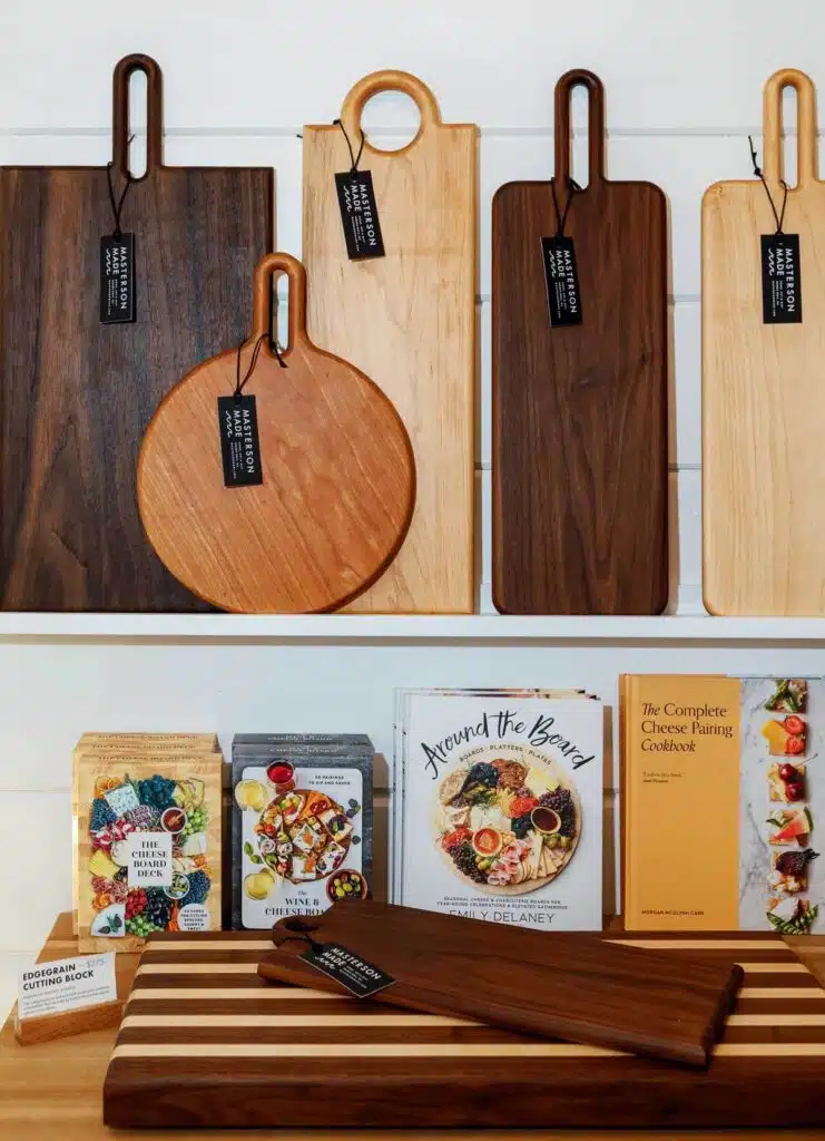 Wooden serving boards hanging on a wall above shelves displaying charcuterie cookbooks and boxed card decks.