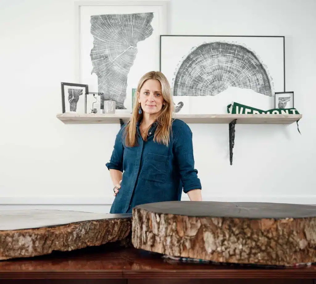 A woman standing behind a table with two large wood cross-section slabs, with framed tree ring art on a shelf behind her.