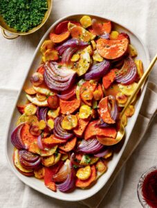 Cider-Glazed Roasted Root Vegetables. Roasted sweet potatoes, yellow carrots, and red onions on a platter with a serving spoon.