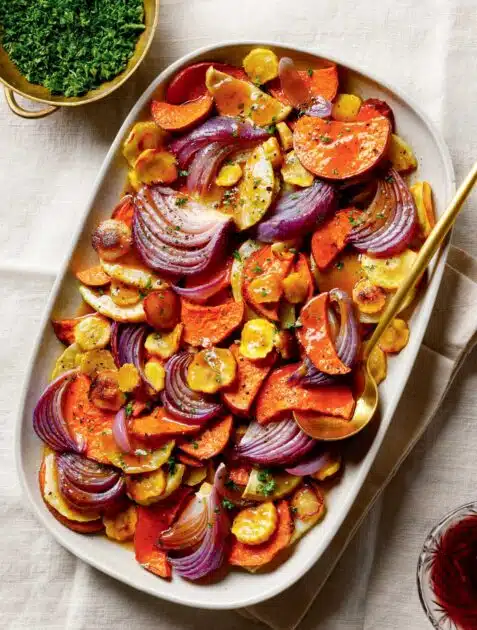 Cider-Glazed Roasted Root Vegetables. Roasted sweet potatoes, yellow carrots, and red onions on a platter with a serving spoon.