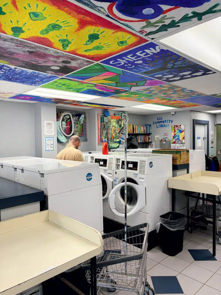 A person using a washing machine in a room with hand-painted ceiling tiles and a small library shelf.