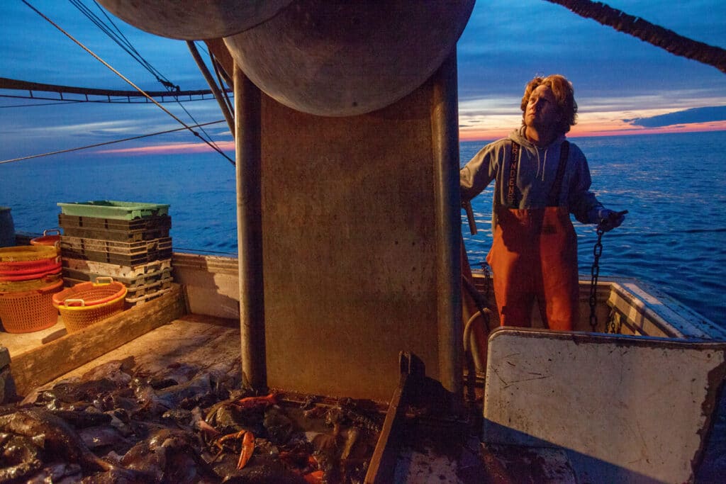 A fisherman in orange overalls works on the deck of a boat at sunset.