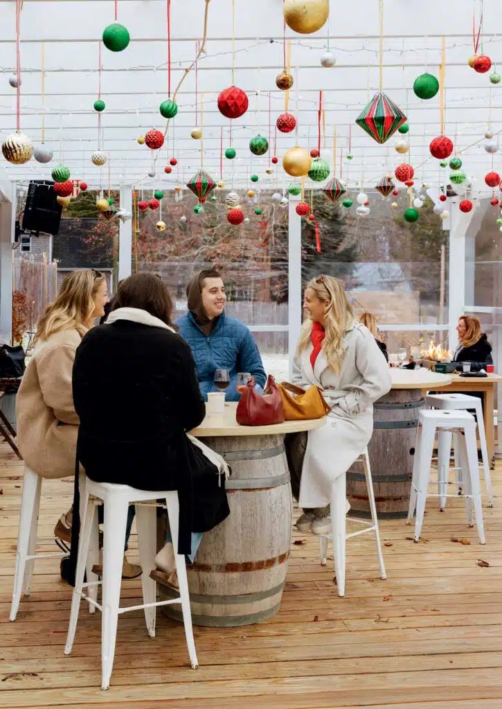 Four people sitting at a wine barrel table on a patio decorated with hanging Christmas ornaments.