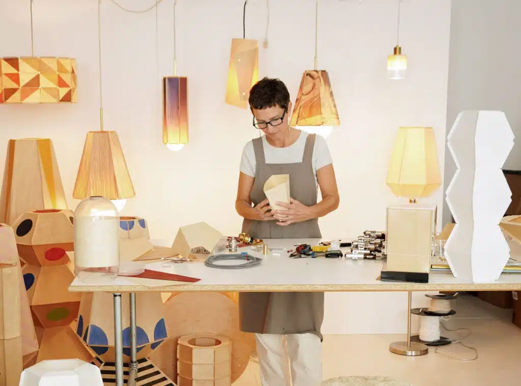 A craftsperson in a studio assembles a wooden lampshade, surrounded by various finished light fixtures.