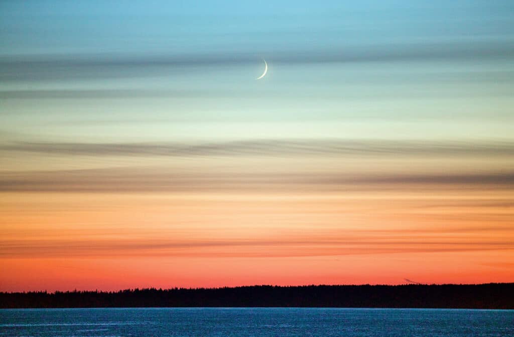 A thin crescent moon hangs in the twilight sky above a dark forest and calm water.