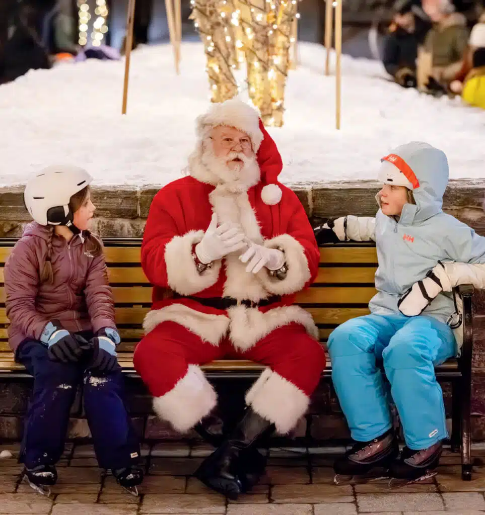 Santa Claus sits on a bench talking to two children in winter clothing and ice skates.