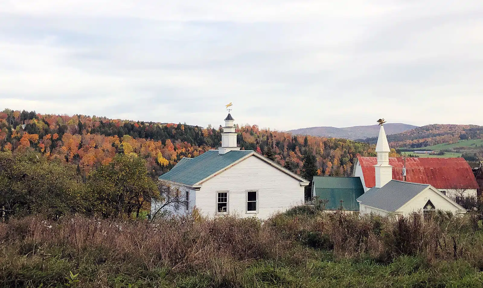 White buildings with steeples nestled before a hillside of autumn foliage.