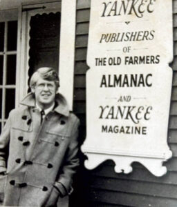 A man in a coat standing next to a sign for Yankee Publishers of The Old Farmer’s Almanac and Yankee Magazine.
