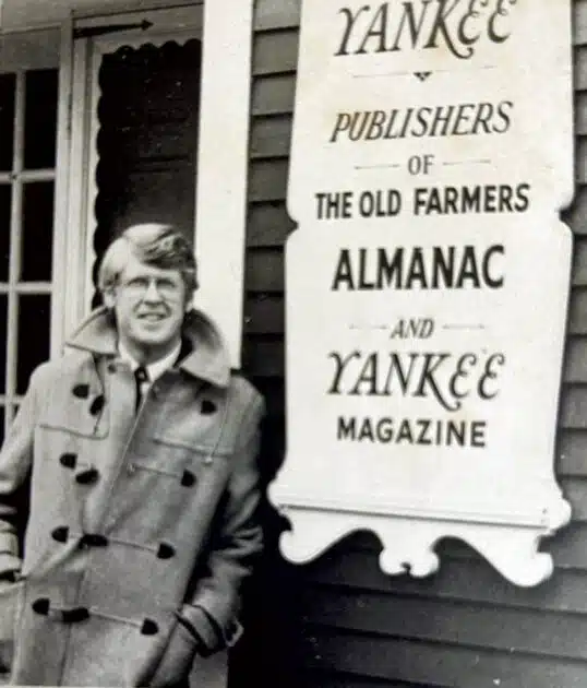 A man in a coat standing next to a sign for Yankee Publishers of The Old Farmer’s Almanac and Yankee Magazine.