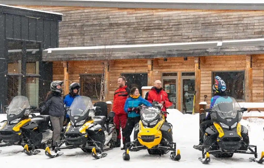 A group of people in winter gear standing with their snowmobiles in front of a wooden building while it snows.