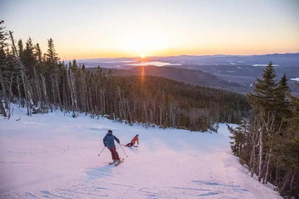 Two skiers descend a snowy slope at sunset, with trees and distant hills in the background.