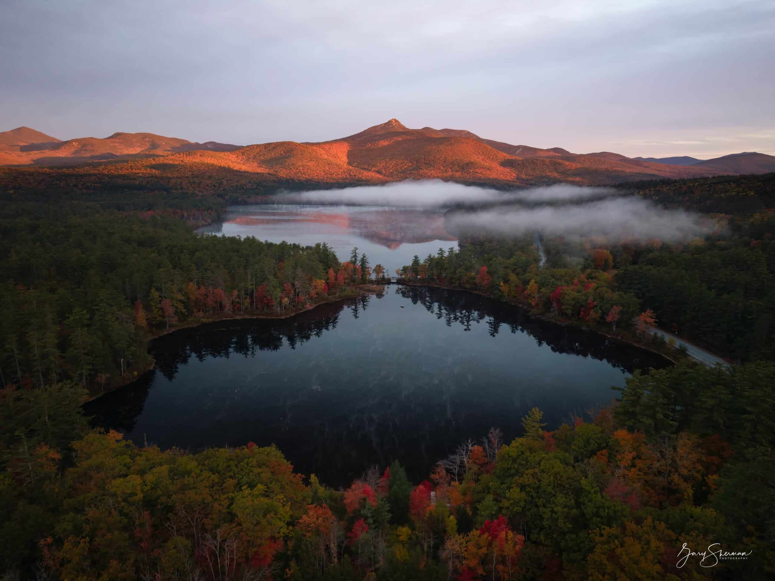 Aerial view of a lake reflecting mountains bathed in warm morning light, surrounded by colorful autumn foliage.