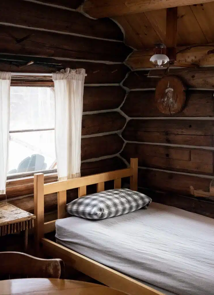 Wooden cabin bedroom with a single bed, checkered pillow, window with curtains, and rustic wooden walls at Little Lyford Lodge & Cabins.
