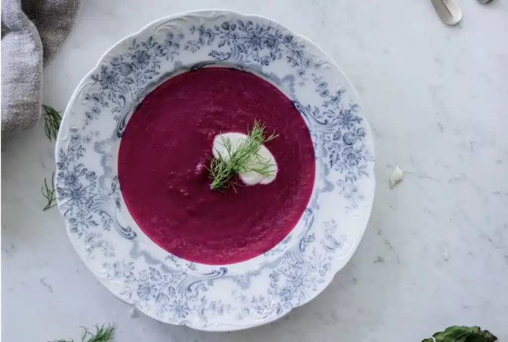 A bowl of beet fennel pear soup with a dollop of cream and dill, served on a blue and white patterned plate.