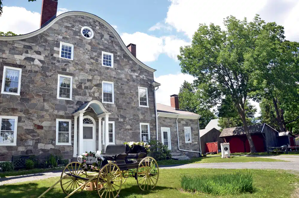 Stone colonial house with arched roof, black carriage with flowers, green lawn, trees, and red barns; site for romantic history in New England