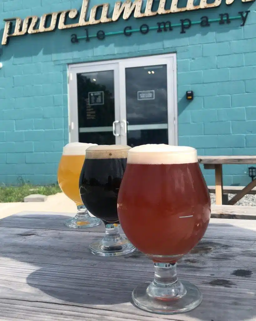 Three different beers in tulip glasses on a picnic table outside a blue brewery building.