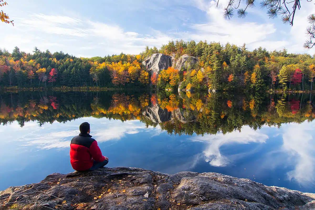 Person in red jacket sits on a rock by a calm lake with fall trees and cliffs reflected in the water. fall foliage planning