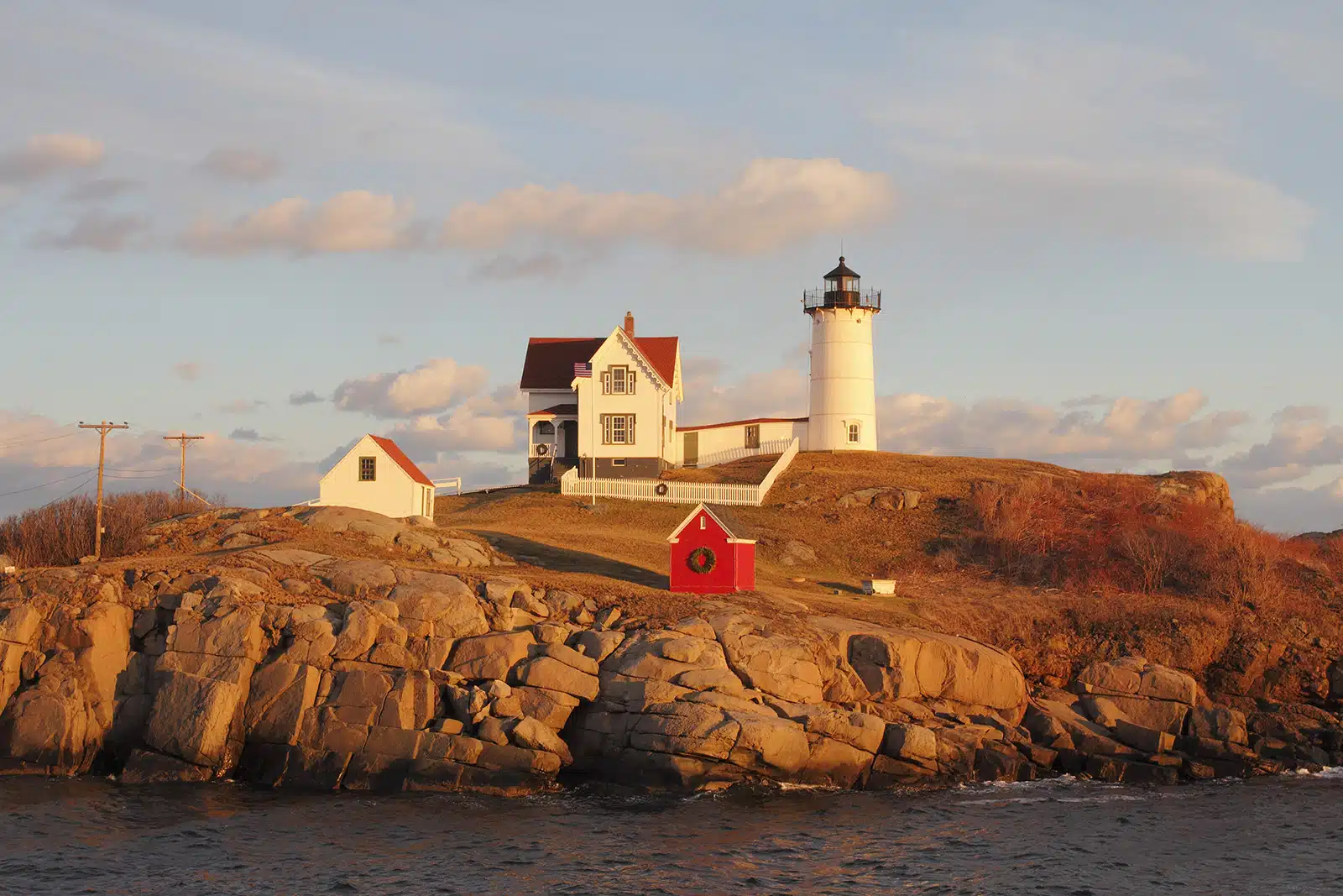 Title: Nubble Lighthouse in December Location: York, ME Judge's comments: "Without the wreath on the red shed, this photograph would be just another Maine landscape image. The artist and the tenants of the property imbue a bit of levity into the scene as the green wreath, meant to greet the passing boat traffic, holds our attention and is meant to greet the passing boat traffic. The white picket fence, two other red roofs and the lighthouse keep our focus within the central cluster. We notice two other small wreaths deliberately placed to lead us down a path to the larger wreath. We enter and leave the photograph through the line of top-left clouds. The long shadows and holiday wreath tip us off that the photo is taken at end of day during early winter."
