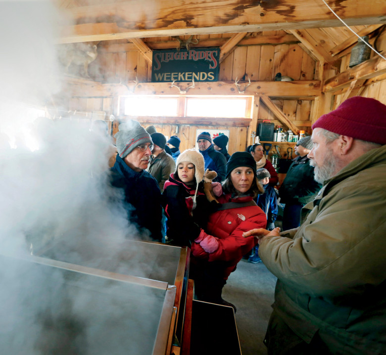 At Jo’s Sugarhouse in Gorham, Maine, volunteer Jim Hanscom chats with the Altman family: Jeremy, daughter Irit, and granddaughter Zahra (peering into the steaming evaporator).