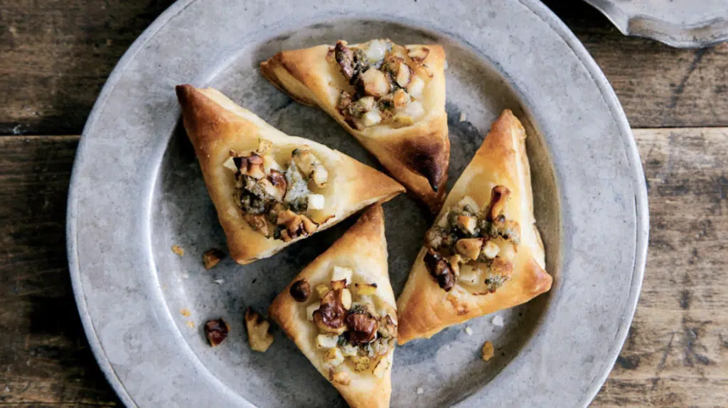 Pear Walnut and Gorgonzola Puffs: Four triangular pastries with nut and herb filling on a round metal plate, on a rustic wooden table.