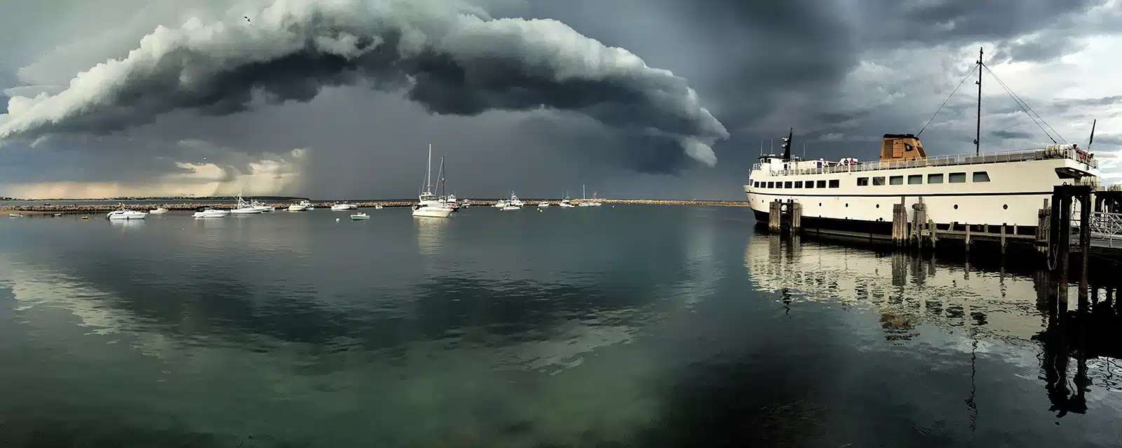 Title: "Storm Over the Old Harbor Location: Block Island, RI Judge's comments: "I am drawn to imagery that is crafted well, gives a fresh and original perspective and evokes narrative inquiry. The artist of “Storm Over the Old Harbor” gives the viewer all three. He uses composition to direct the central focus to the storm in a number of ways. The ship draws the viewer in, as if pointing towards danger. The cloud and its reflection, with the centralized white boat and mast form the eye of potential activity. The photograph itself is not cliché and leaves us to linger and imagine the meaning of the moment caught by the artist."