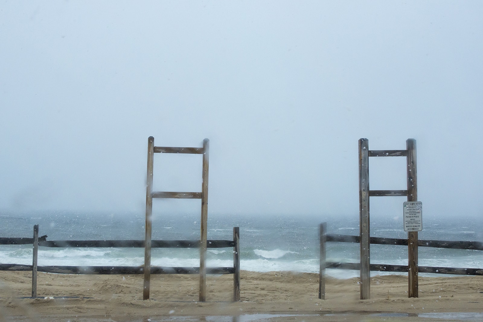 Title: Snow Location: Truro, MA Judge's comments: "Weather is a constant in New England, from the coastline beaches to its mountain heights. Any photographer in search of landscape drama has to learn to withstand weather and the elements of heat and cold. The photographer who took this photograph stood on a windswept beach to capture weather in process. There is sleet on the lens and blurred imagery that suggests wind with snow. The palette contains muted blues and beiges that are turning towards gray. The geometry of the horizontal fencing mixes with the vertical lines of the signage, holding back entrance to the planes of sand, sea and sky despite the Coast Guard’s message that the “Water Quality is Acceptable.”" 