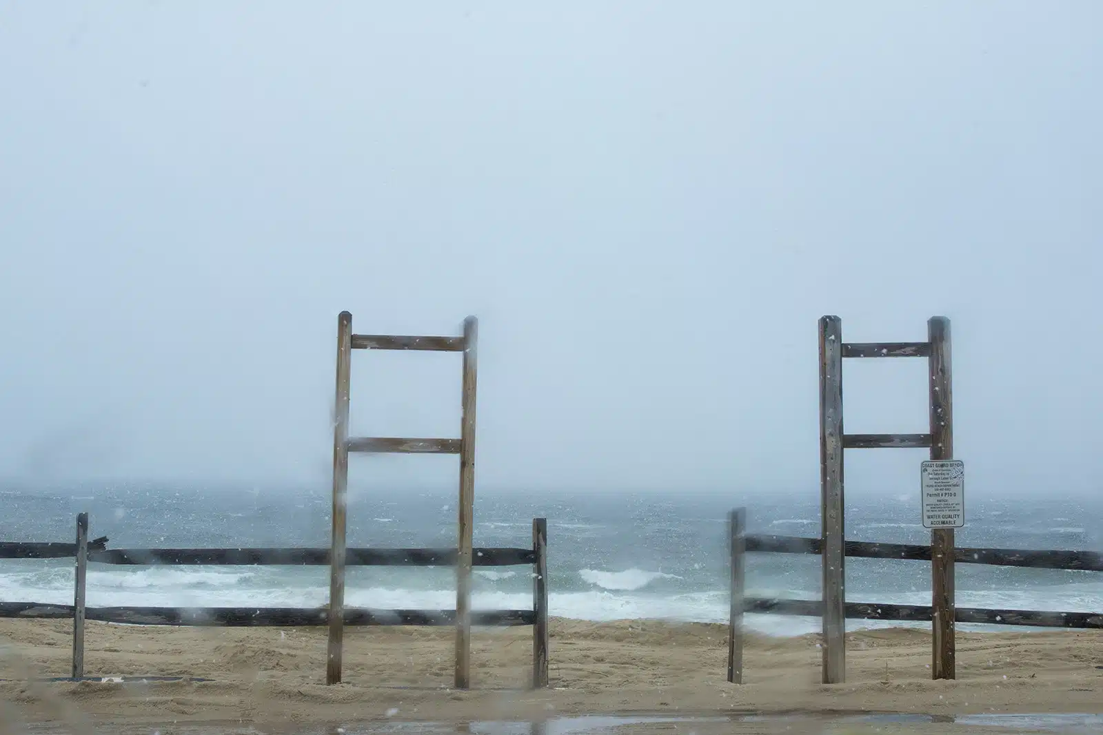 Title: Snow Location: Truro, MA Judge's comments: "Weather is a constant in New England, from the coastline beaches to its mountain heights. Any photographer in search of landscape drama has to learn to withstand weather and the elements of heat and cold. The photographer who took this photograph stood on a windswept beach to capture weather in process. There is sleet on the lens and blurred imagery that suggests wind with snow. The palette contains muted blues and beiges that are turning towards gray. The geometry of the horizontal fencing mixes with the vertical lines of the signage, holding back entrance to the planes of sand, sea and sky despite the Coast Guard’s message that the “Water Quality is Acceptable.”"