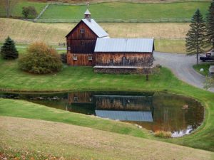Small wooden barn with a tin roof next to a pond in a rural landscape, surrounded by rolling green hills, trees, and a winding dirt road.