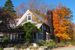 A rustic house with stone siding and a chimney, surrounded by vibrant autumn trees with brightly colored leaves under a clear blue sky.